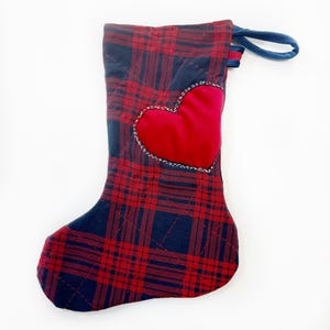 May include: A Christmas stocking in a red and navy plaid design. A red heart is appliquéd on the front, edged with white stitching. It features a blue hanging loop.