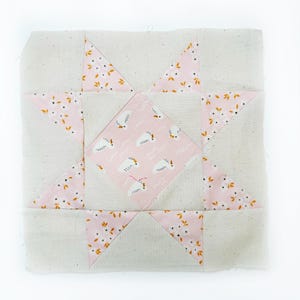 May include: A pink and white patchwork quilt block with a star design. The star is made of pink fabric with a white floral pattern. The background is a light beige fabric.