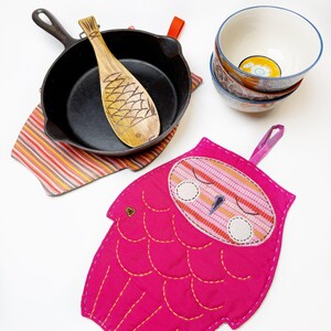 May include: A pink owl-shaped pot holder with a striped design on the face. The pot holder is made of fabric and has a loop for hanging.