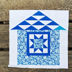 May include: A square quilt block featuring a house design. The roof is made of blue triangles, and the house body has a floral pattern in shades of blue. A white border surrounds the house shape.