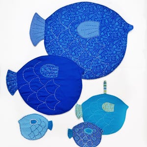 Puede incluir: Una colección de seis peces de tela azul de varios tamaños. Los peces tienen patrones detallados, incluyendo diseños florales y de puntos, con costuras blancas y aletas contrastantes. La paleta de colores varía de azul claro a oscuro.
