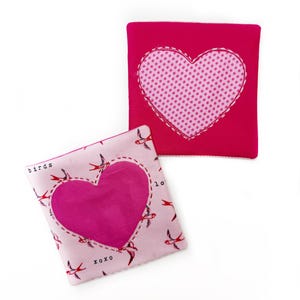 May include: Two pink fabric heart-shaped pouches. One pouch has a pink polka dot heart sewn on the front. The other pouch has a solid pink heart sewn on the front and a pattern of red birds with the text "birds" and "xoxo" on the front.