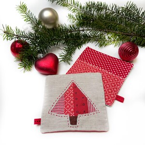 Könnte beinhalten: Zwei rote und weiße Topflappen in Weihnachtsbaumform mit Leinenhintergrund. Die Topflappen sind von einem Tannenzweig und roten und silbernen Ornamenten umgeben.