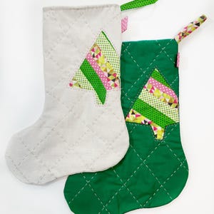 Könnte beinhalten: Zwei Weihnachtsstrümpfe, einer weiß und einer grün, mit einem gesteppten Rautenmuster. Beide Strümpfe haben ein Patchwork-Weihnachtsbaumdesign mit grünem, rosa und gelbem Stoff.