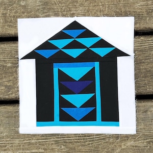 May include: A square fabric block featuring a house design. The roof is black with blue triangles, and the body is black with blue and teal accents. The block is set against a white background.
