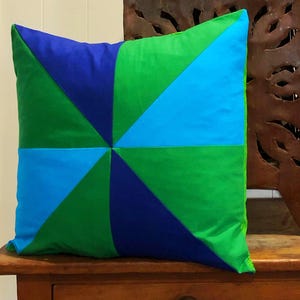 May include: A square cushion with a geometric pattern of blue, green, and turquoise triangles. The cushion is made of fabric and has a white border.