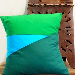 May include: A square decorative pillow with a color block design. The pillow features three sections: a top section of bright green, a middle section of turquoise blue, and a bottom section of dark green. The pillow is made of a soft fabric.