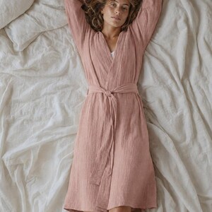 May include: A dusty rose-colored robe featuring a textured, crinkled fabric finish, long sleeves, and a matching fabric tie belt at the waist. The garment has a V-neckline and falls to a knee-length hem, providing a relaxed, lightweight appearance.