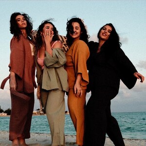 May include: Four women wearing different colored pantsuits stand on a beach. The women are smiling and laughing. The pantsuits are all in earth tones and have a relaxed, flowy fit.