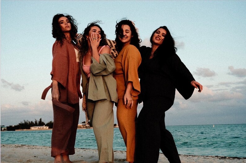May include: Four women wearing different colored pantsuits stand on a beach. The women are smiling and laughing. The pantsuits are all in earth tones and have a relaxed, flowy fit.