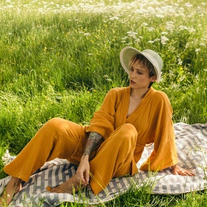 May include: A person wearing a mustard yellow jumpsuit and a light green straw hat sits on a white and gray striped blanket in a field of green grass and white flowers.