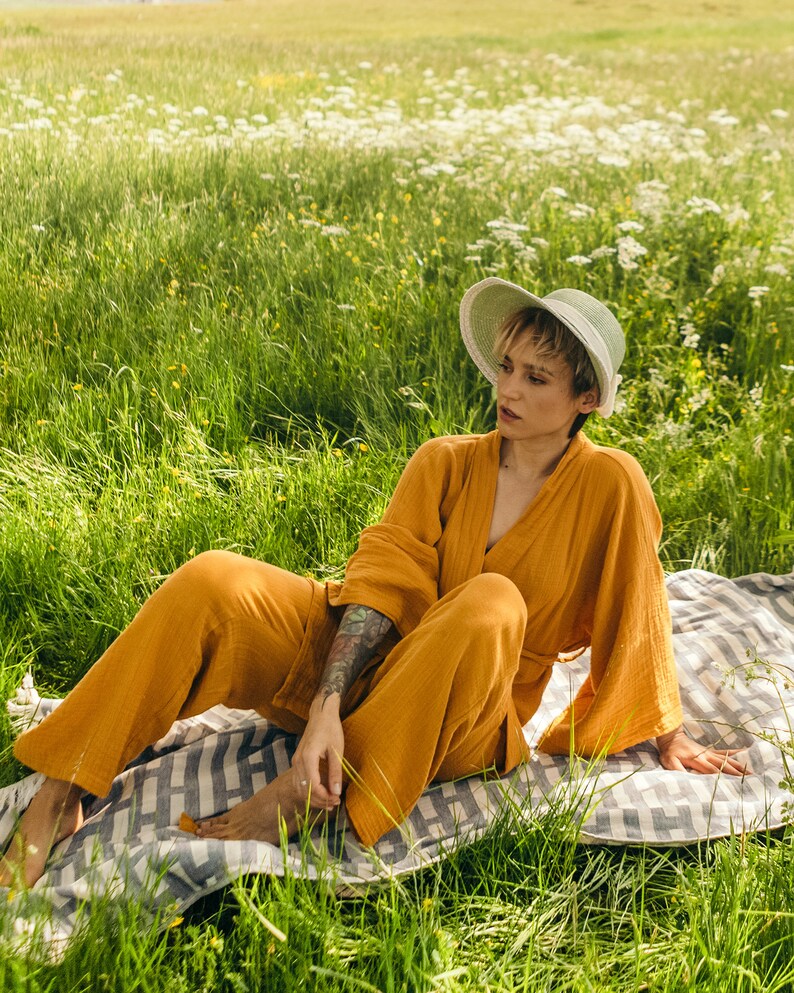 May include: A person wearing a mustard yellow jumpsuit and a light green straw hat sits on a white and gray striped blanket in a field of green grass and white flowers.