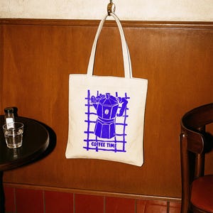 Peut inclure: Sac fourre-tout crème avec un motif bleu d'une cafetière et l'inscription "COFFEE TIME". Le sac est suspendu à un crochet en métal. L'arrière-plan comprend un mur en bois, une table et une chaise.