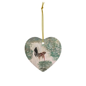 May include: Heart-shaped ornament featuring a watercolor painting of a person walking a dog in a snowy forest. The ornament has a gold string for hanging. The artwork is in shades of green, brown, and white.