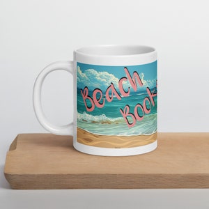 Peut inclure: Une tasse en céramique blanche avec une scène de plage et le texte "Beach Book" en lettres roses.