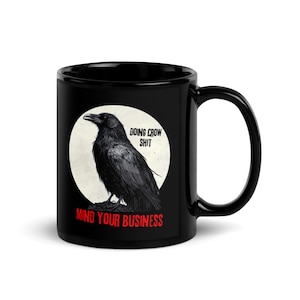 May include: Black ceramic mug featuring a detailed illustration of a crow inside a white circle. The mug has the text "DOING CROW SHIT" and "MIND YOUR BUSINESS" in red. The mug has a large handle.