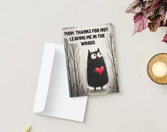 Puede incluir: Una tarjeta de felicitación con un gato negro sosteniendo un corazón rojo, con el texto "MOM, THANKS FOR NOT LEAVING ME IN THE WOODS." La tarjeta está en un sobre blanco. Una vela encendida en un portavelas dorado está al lado de la tarjeta.