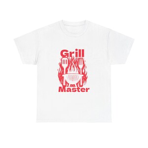 May include: White t-shirt with a red graphic design. The design features the words "Grill Master" above an illustration of a grill, spatula, and fork, all in red. The design is surrounded by a flame-like pattern.