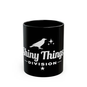 Puede incluir: Taza de cerámica negra con el texto blanco "Shiny Things Division", una ilustración de cuervo blanco y estrellas. La taza es cilíndrica y tiene un acabado brillante.