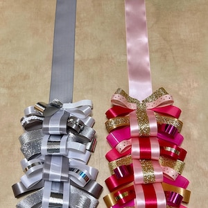 May include: Two decorative ribbon cascades featuring loops of various textures and finishes. The left cascade uses silver, white, and grey ribbons with metallic and sequin accents. The right cascade features pink, red, and gold glitter ribbons.