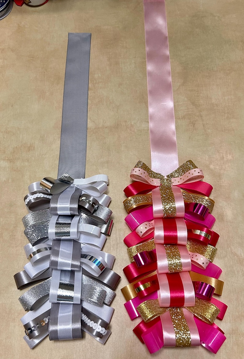 May include: Two decorative ribbon cascades featuring loops of various textures and finishes. The left cascade uses silver, white, and grey ribbons with metallic and sequin accents. The right cascade features pink, red, and gold glitter ribbons.