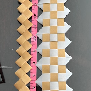 May include: Two woven ribbon strips placed next to a pink measuring tape. The left strip is solid gold, while the right strip features a checkered pattern of gold and translucent white. Both strips are secured with metal staples at the ends.