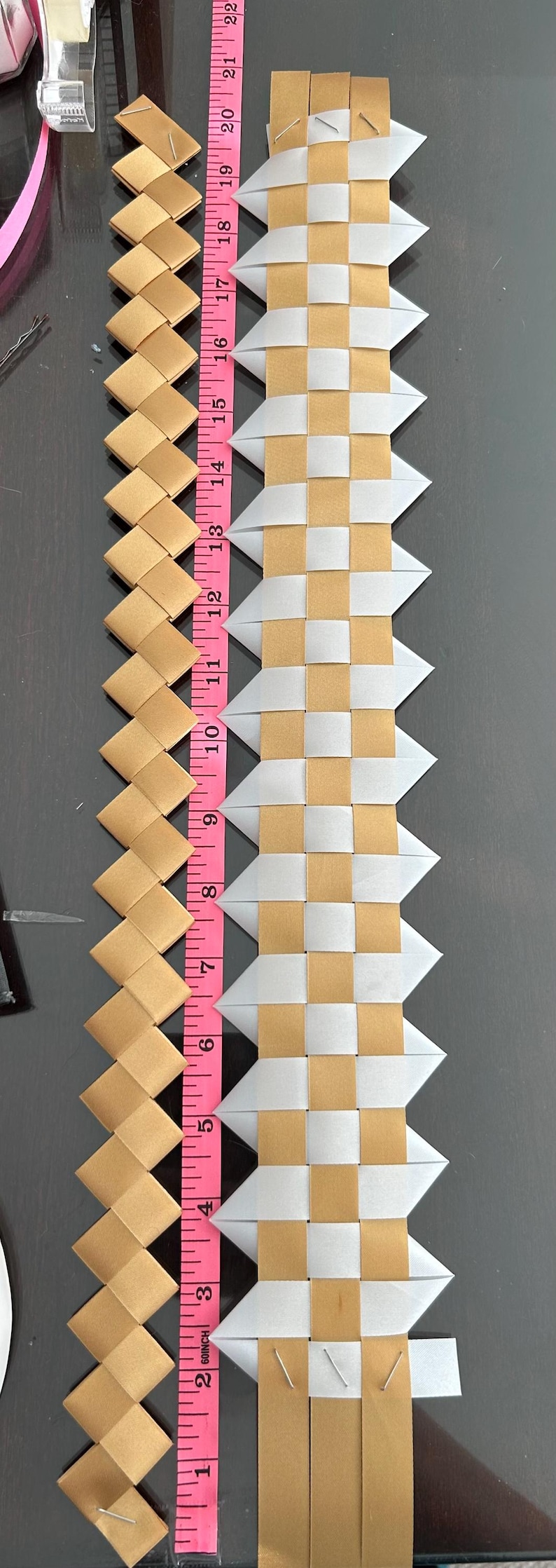 May include: Two woven ribbon strips placed next to a pink measuring tape. The left strip is solid gold, while the right strip features a checkered pattern of gold and translucent white. Both strips are secured with metal staples at the ends.
