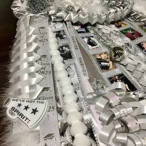 May include: A white and silver homecoming mum featuring layered ribbons, white faux flowers, and pom-pom trim. The ribbons display the text Class of 2025, Senior, and a megaphone cutout with the text WE'VE GOT THE SPIRIT! along with several small photos.