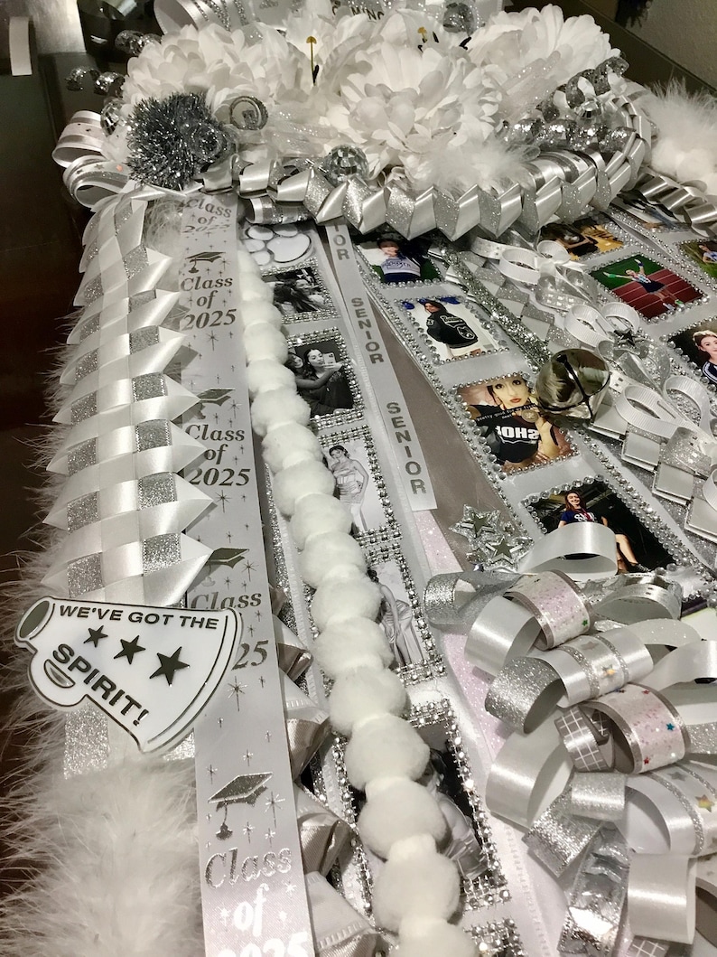 May include: A white and silver homecoming mum featuring layered ribbons, white faux flowers, and pom-pom trim. The ribbons display the text Class of 2025, Senior, and a megaphone cutout with the text WE'VE GOT THE SPIRIT! along with several small photos.