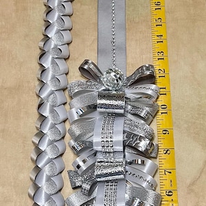 May include: A silver and white homecoming mum accessory featuring a long braided ribbon strand and a decorative cluster of layered ribbons, metallic accents, and a disco ball bead, measured by a yellow tape measure reaching 25 inches in length.