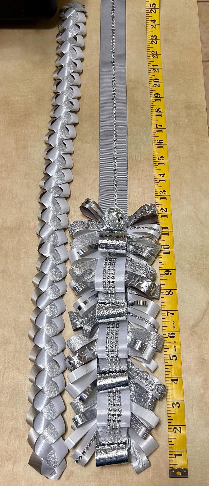 May include: A silver and white homecoming mum accessory featuring a long braided ribbon strand and a decorative cluster of layered ribbons, metallic accents, and a disco ball bead, measured by a yellow tape measure reaching 25 inches in length.