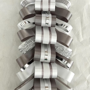 May include: A decorative ribbon ornament featuring a vertical arrangement of layered loops in shades of white, silver, and grey. The design incorporates various textures, including smooth satin, metallic silver accents, and intricate silver lace trim.
