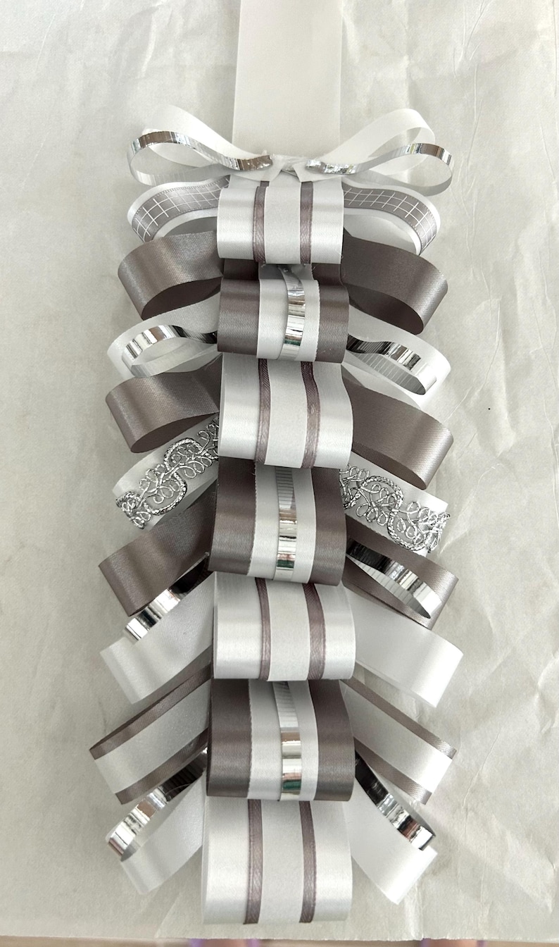 May include: A decorative ribbon ornament featuring a vertical arrangement of layered loops in shades of white, silver, and grey. The design incorporates various textures, including smooth satin, metallic silver accents, and intricate silver lace trim.