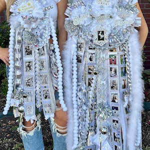 May include: Two large white and silver homecoming mums featuring layered ribbons, white faux fur trim, metallic silver accents, and multiple small square photo prints. The designs include decorative lettering, small charms, and glittery embellishments.