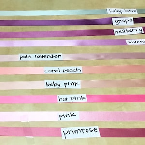 May include: A collection of ten horizontal satin ribbons in various colors, each labeled with a white paper tag. The colors include baby blue, grape, mulberry, lavender, pale lavender, coral peach, baby pink, hot pink, pink, and primrose.