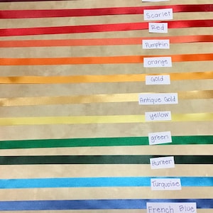 May include: A collection of horizontal satin ribbons in various colors, each labeled with a white paper tag. The colors include purple, cyclamen, burgundy, scarlet, red, pumpkin, orange, gold, antique gold, yellow, green, hunter, turquoise, French blue, royal, and navy.