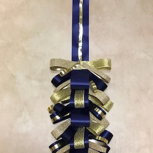 May include: A decorative hanging ornament constructed from layered loops of navy blue satin ribbon and gold glitter ribbon. The design features a long vertical navy blue ribbon strip at the top, with alternating gold and blue ribbon loops arranged in a cascading, symmetrical pattern that tapers toward the bottom.