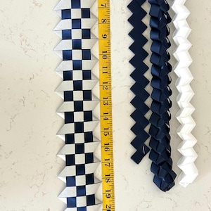 May include: Four woven ribbon leis, one featuring a silver and navy blue checkered pattern measuring 26 inches long, two solid navy blue braided ribbons, and one solid white braided ribbon, all displayed alongside a yellow measuring tape.