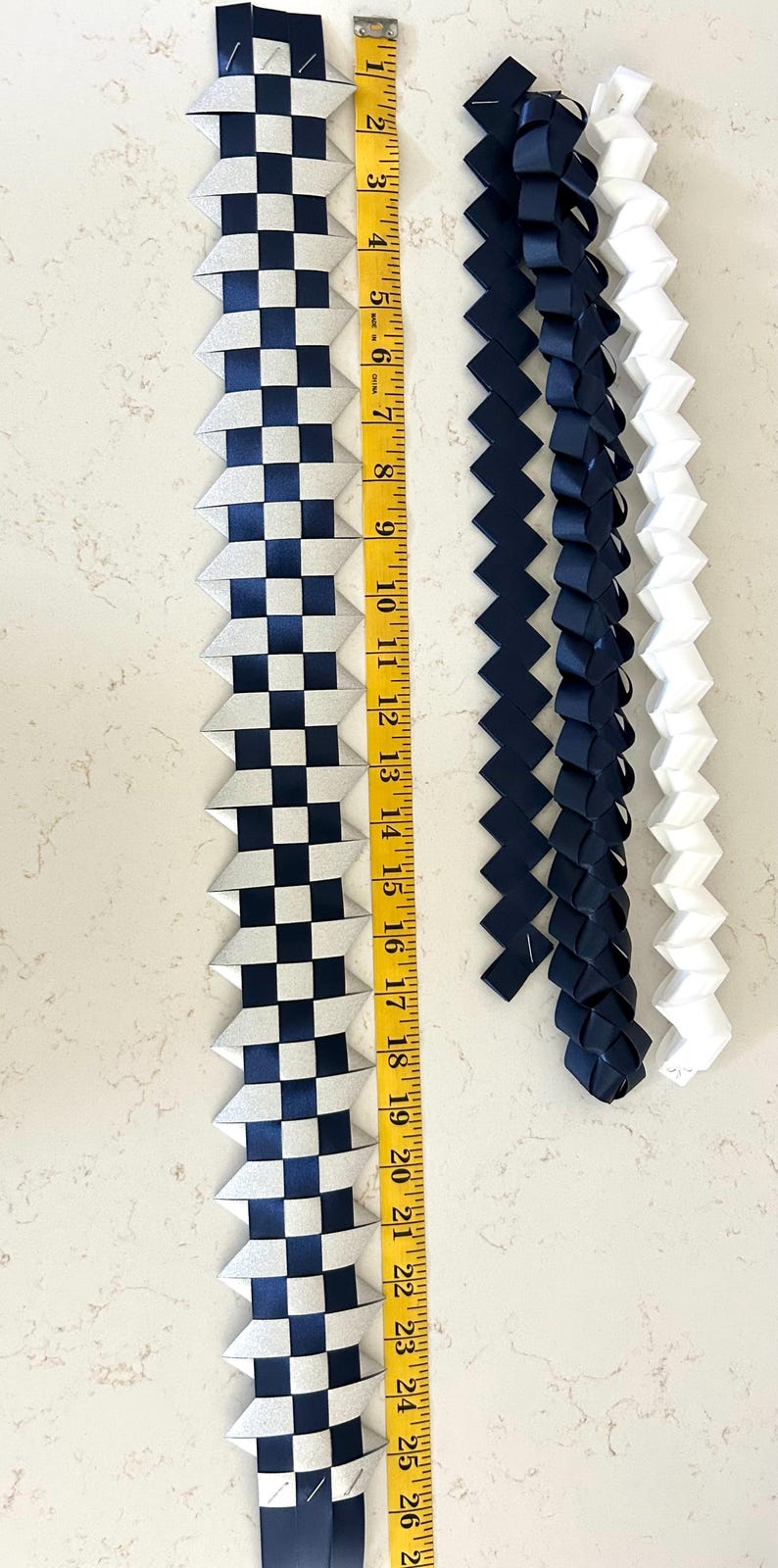 May include: Four woven ribbon leis, one featuring a silver and navy blue checkered pattern measuring 26 inches long, two solid navy blue braided ribbons, and one solid white braided ribbon, all displayed alongside a yellow measuring tape.