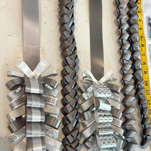 May include: Four grey and silver ribbon homecoming mums and braided ribbon strands. Two mums feature layered loops of solid, glittered, and patterned silver and grey ribbons with white bows. Two braided ribbon strands are included, with a yellow ruler showing 13 inches.