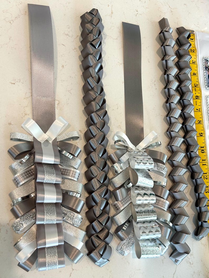 May include: Four grey and silver ribbon homecoming mums and braided ribbon strands. Two mums feature layered loops of solid, glittered, and patterned silver and grey ribbons with white bows. Two braided ribbon strands are included, with a yellow ruler showing 13 inches.