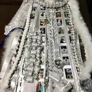 May include: A white and silver homecoming mum featuring numerous ribbons, white artificial flowers, and small square photographs. The ribbons are printed with Class of 2025, Senior, and Spirit text. It includes silver bells and white feather trim.