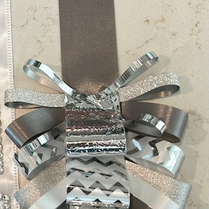 May include: A decorative ribbon ornament featuring a long taupe satin strap attached to a layered cluster of ribbons. The cluster includes silver glitter, metallic silver chevron-patterned, and white lace-trimmed ribbons arranged in loops.