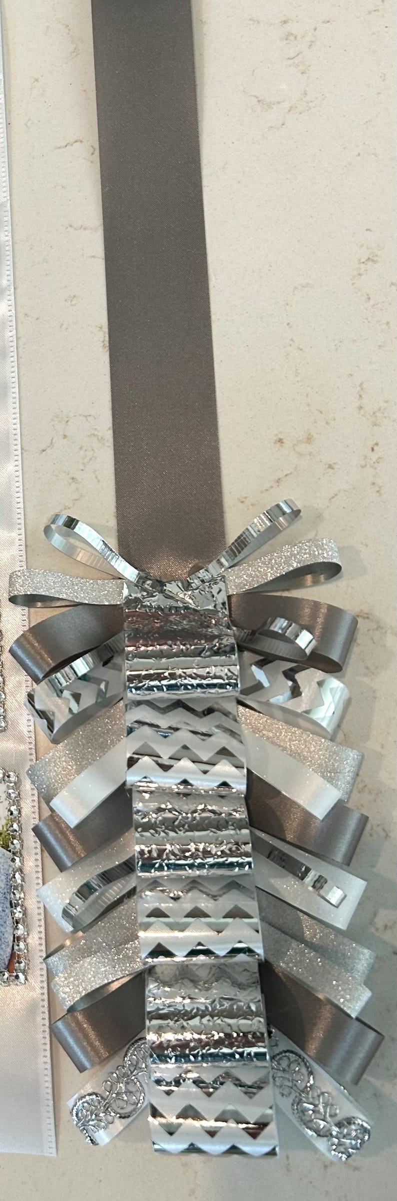 May include: A decorative ribbon ornament featuring a long taupe satin strap attached to a layered cluster of ribbons. The cluster includes silver glitter, metallic silver chevron-patterned, and white lace-trimmed ribbons arranged in loops.