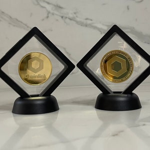 May include: Two gold-coloured Chainlink cryptocurrency coins in black display cases with clear lids. The coins feature a hexagonal design with the Chainlink logo and text.