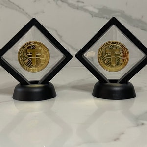 May include: Two gold-coloured cryptocurrency coins with a black 'T' in the centre are displayed in clear diamond-shaped display cases with black bases.