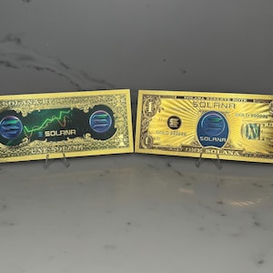 Peut inclure: Deux billets de banque de couleur or avec le mot "Solana" imprimé dessus. Un billet présente un graphique en ligne verte et l'autre billet présente un logo bleu et vert. Les deux billets portent l'inscription "One Solana".