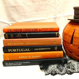 May include: An orange ceramic pumpkin with carved face details and a brown top hat sits next to a stack of books. The books have orange and black covers with titles like "Calrdon Cookery" and "Portugal". A black lace trim is at the base.