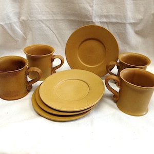 May include: A set of six tan-colored ceramic mugs and saucers. The mugs have curved handles and flared rims. The saucers are round with a slightly raised center. The set is arranged on a white surface.