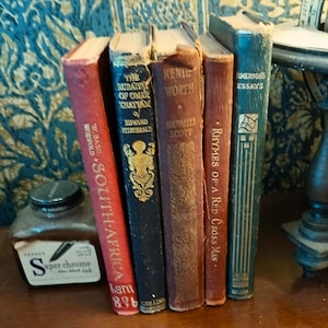 May include: A collection of antique books with worn covers, including titles like "The Rubaiyat of Omar Khayyam" and "Emerson's Essays." The books are arranged together with an ink bottle labeled "Super Chrome" and a decorative table.